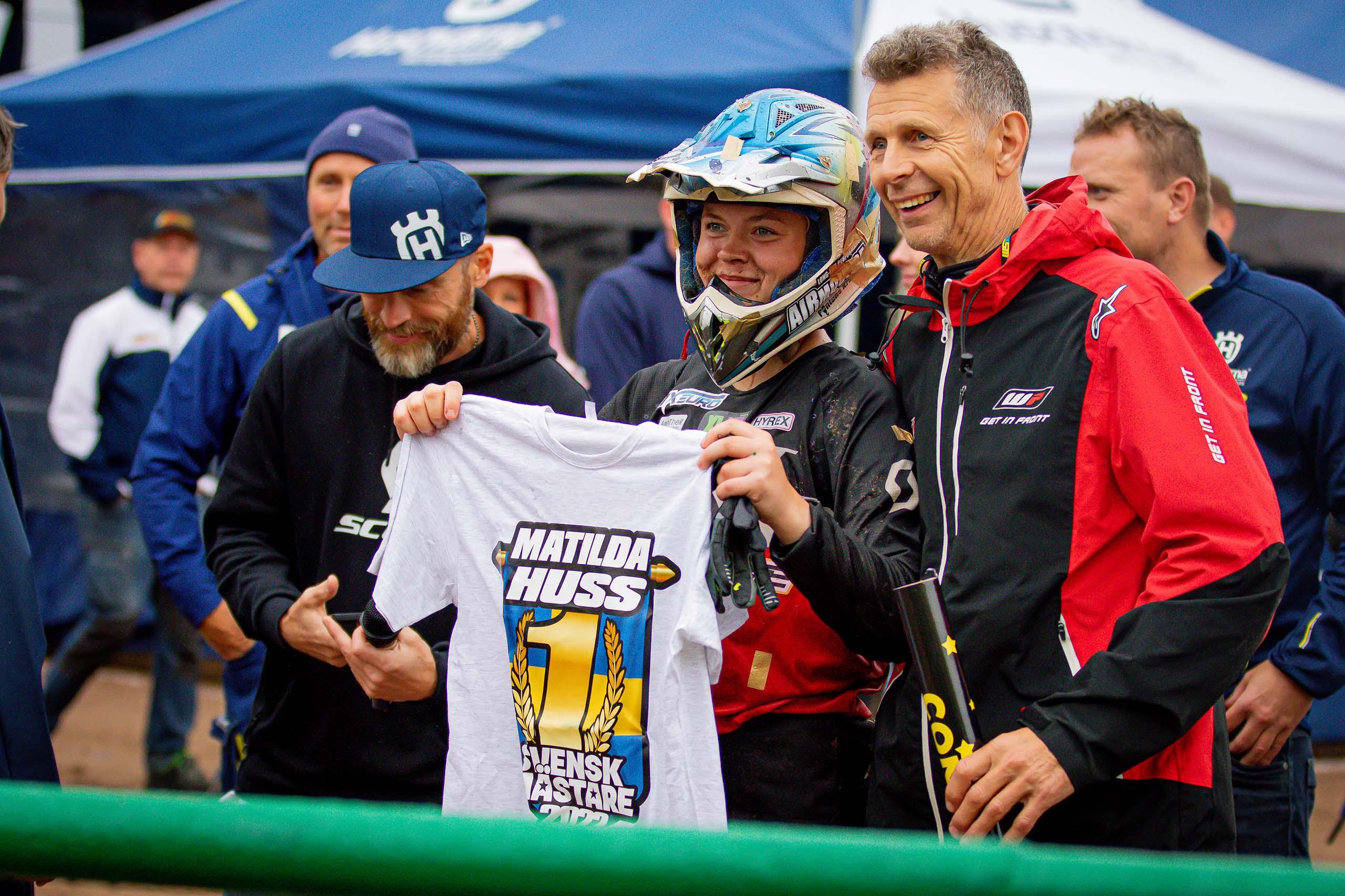 En kvinnlig motocrossförare håller leende upp en t-shirt som det står "Matilda Huss Svensk Mästare 2022" på.